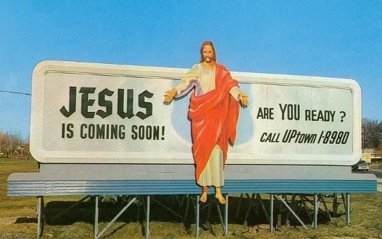 A road sign that reads JESUS IS COMING SOON! ARE YOU READY? CALL UPtown 1-8980, with a cutout picture of a Caucasian Jesus in a white robe and red shroud.